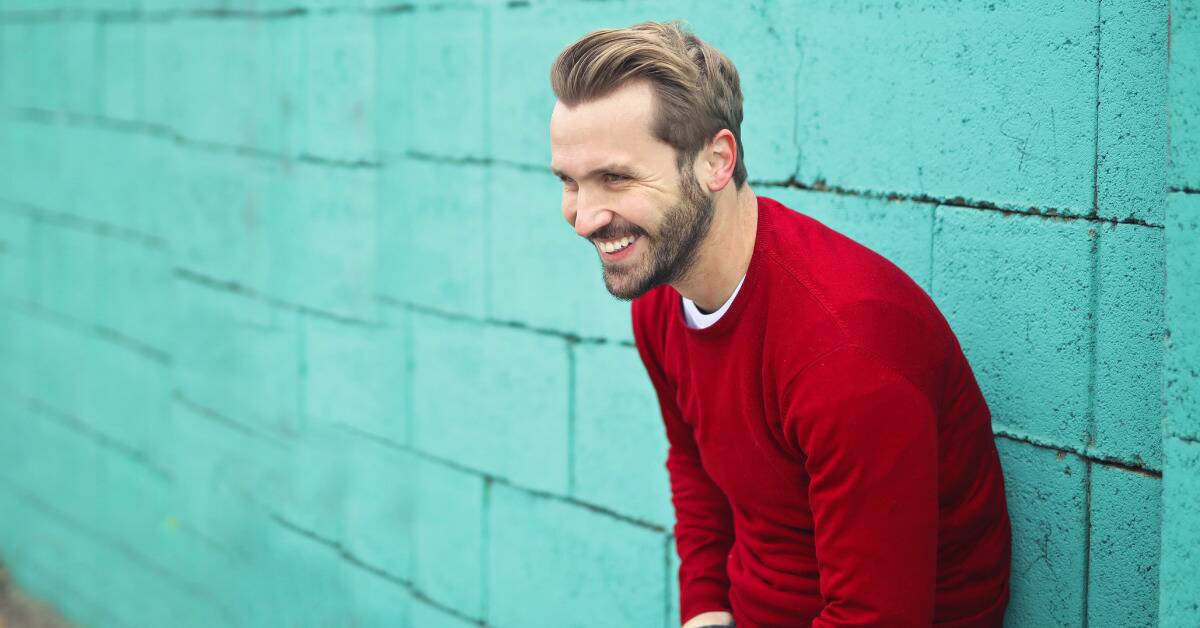 A man in a red sweater smiling as he leans up against a bright teal wall.