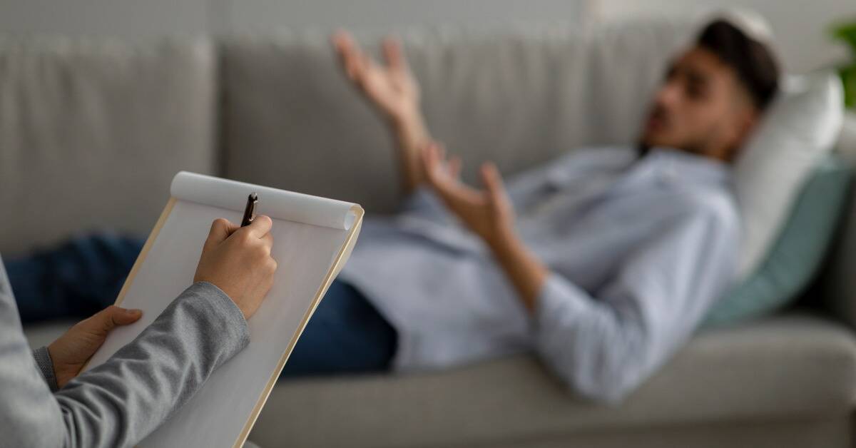 A man laying on a couch in the background, mid-talking, while a therapist in the background gets ready to start writing notes on a notepad.