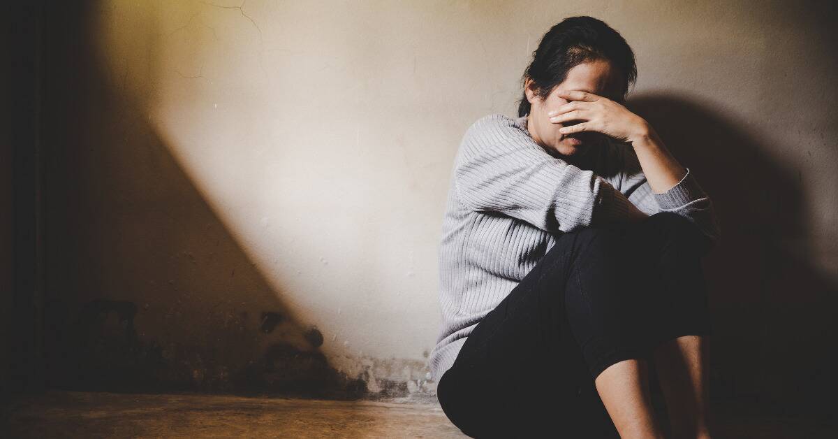 A woman sat on the floor, back against the wall, knees drawn up to her chest, covering her face with one hand.