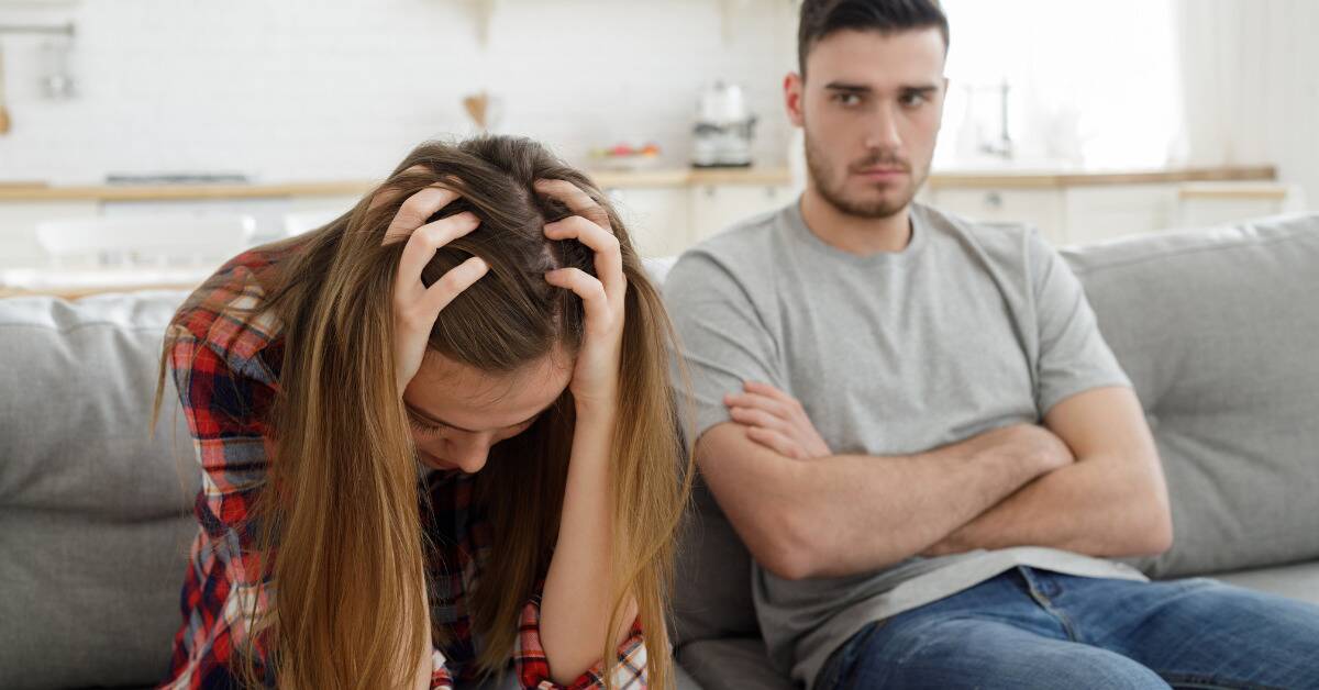 A couple sat on a couch, the woman leaning forward with her head in her hands, while the man sits with his arms crossed, looking at her with an annoyed expression.