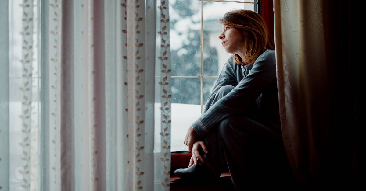 A woman sat on a window ledge, looking forlornly outside at the winter snow.