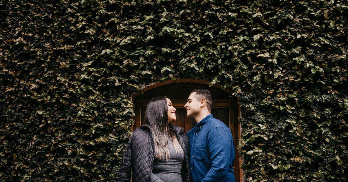 A low shot of a couple smiling at each other in front of a plant-covered wall.