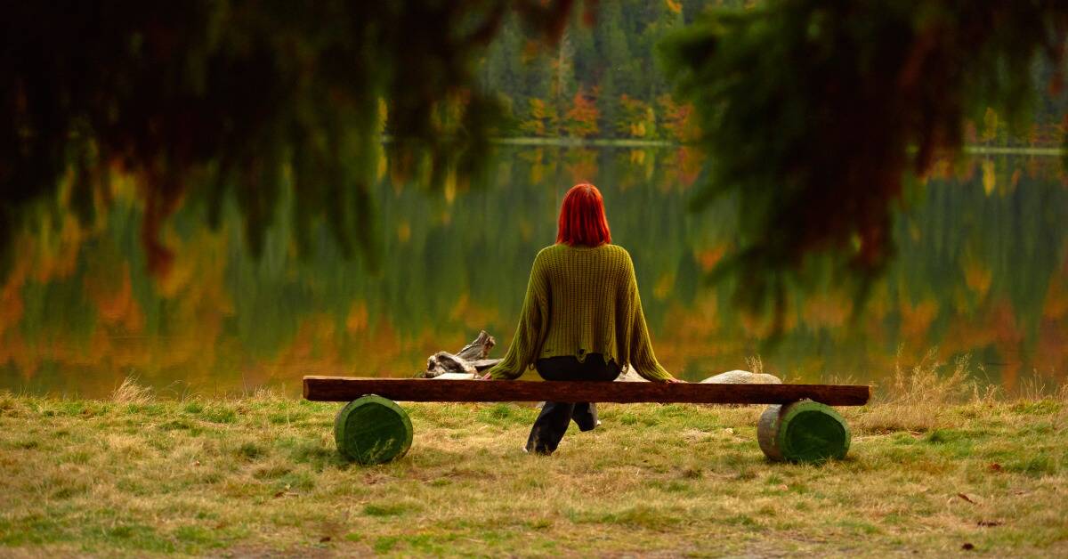 A woman with bright red hair sitting on a bench in front of the water, trees surrounding her.
