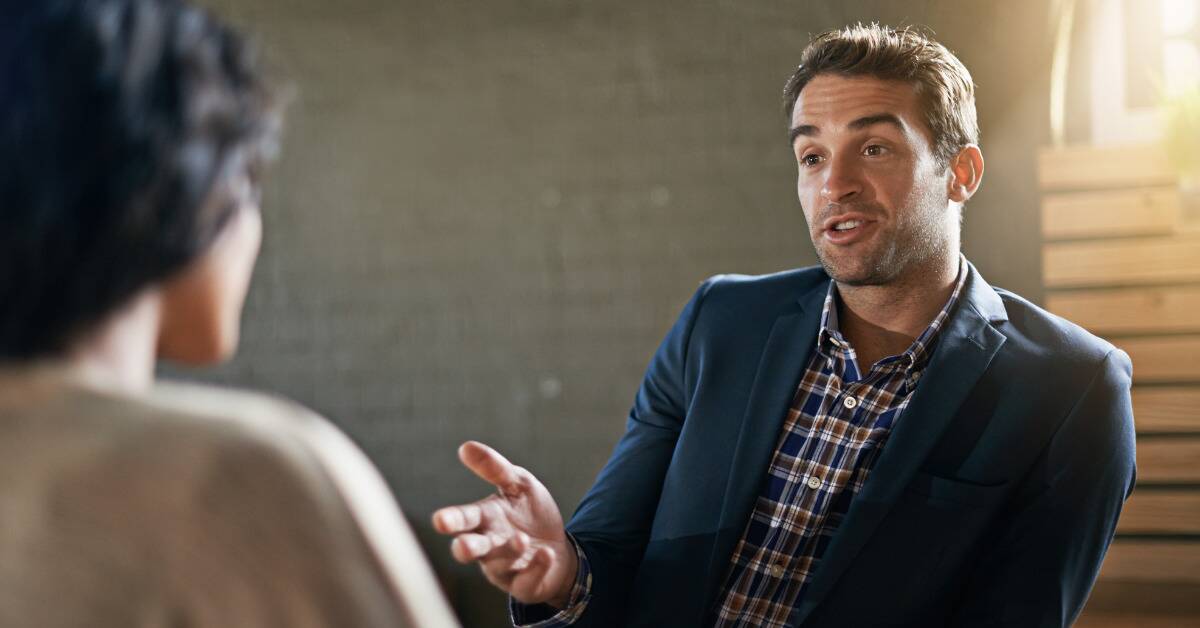 Two people talking, the camera over one person's shoulder to focus on the man speaking, gesturing with his hand. 