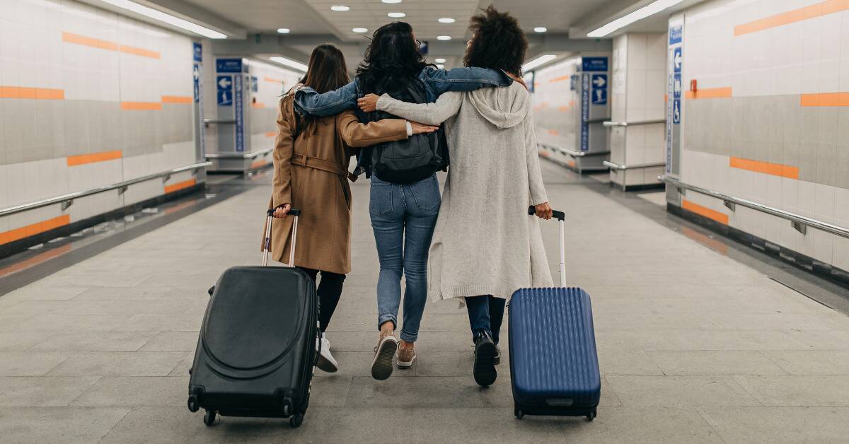 Three friends with their arms around one another two pulling suitcases, as they walk down an airport hallway.