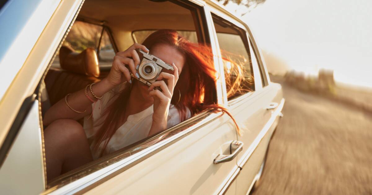 A woman leaning out the window of a car while taking a picture on a film camera.