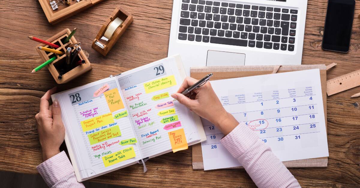 An arial shot of someone working at their desk, laptop open, writing in two copies of their calendars, one daily and one mothly.