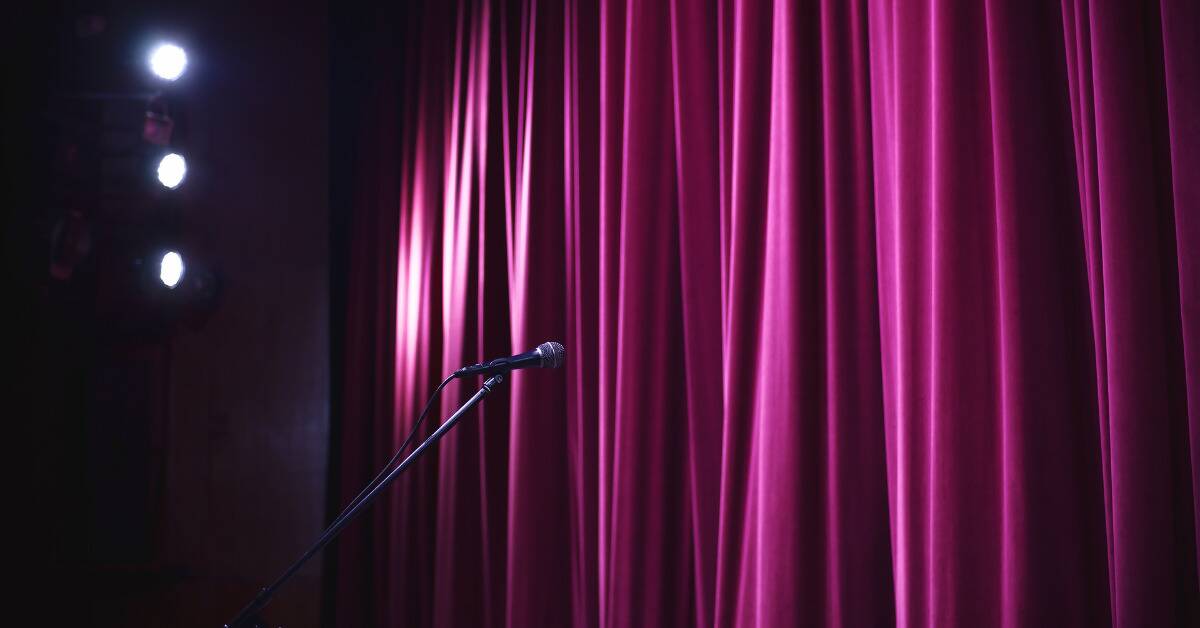 A microphone on a lone stage, in the spotlight, set up in front of purple stage curtains.