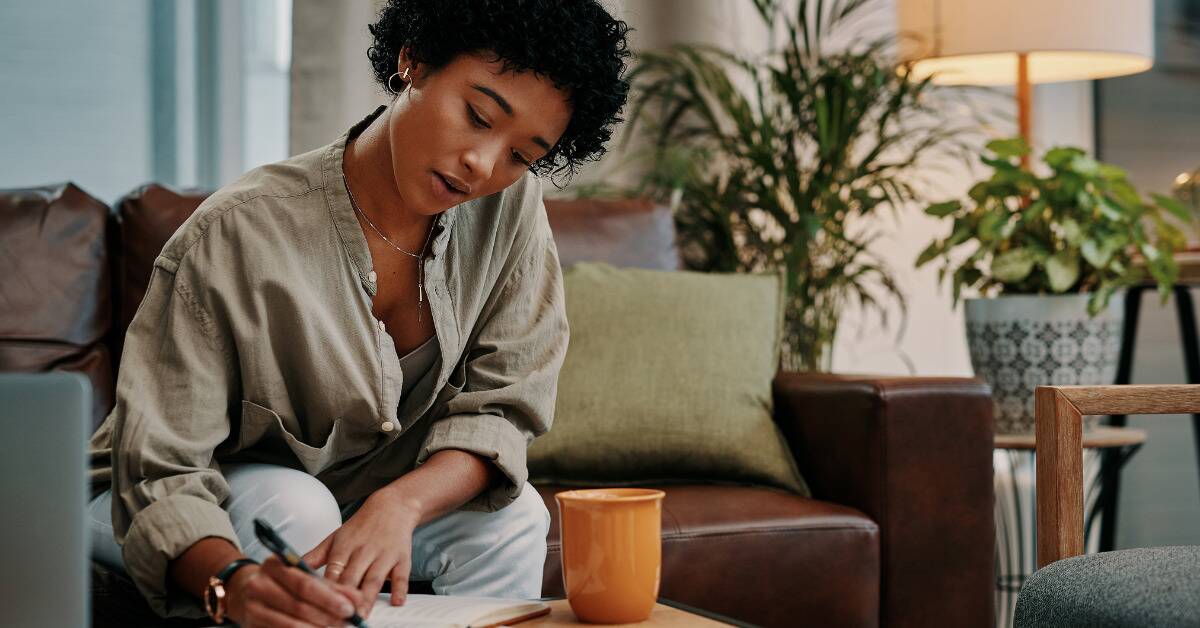 A woman sitting on her couch, leaning forward to write in a journal that's on her coffee table.