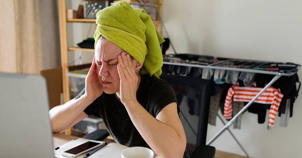 A woman sat at her desk, her hair wrapped in a bright green towel, fingers pressed to her temple as she's clearly stressed.