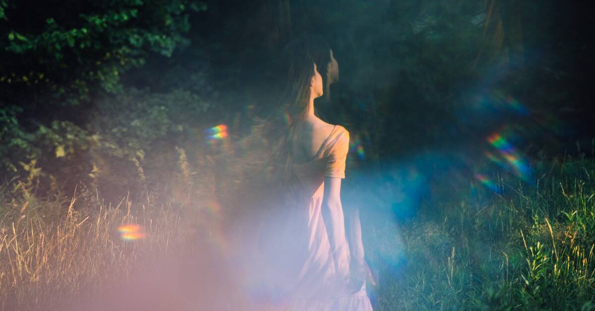 A woman in ethereal rainbow light walking through a field of tall grass.