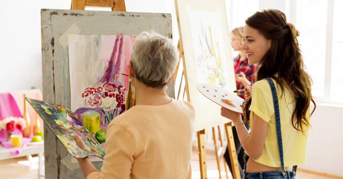An older woman painting in a group setting, a younger woman looking over at her painting and smiling.