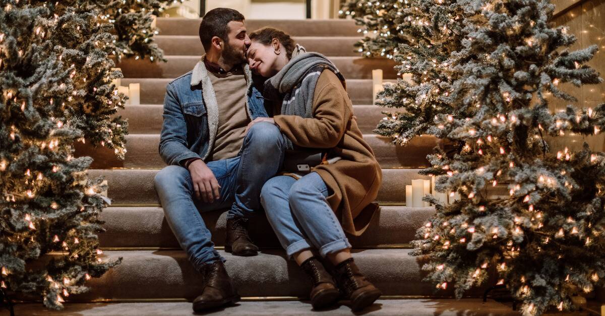 A couple sitting on a stairwell decorated with garland and lights, the woman leaning into the man's side while he kisses her forehead.