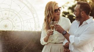 A couple chatting as they cheers two glasses of champagne, free arms linked together, a golden astrology wheel superimposed on the left side of the image.