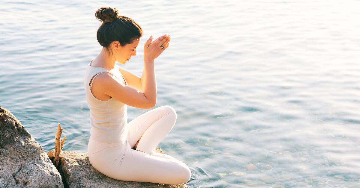 A woman sitting on a rock next to the water, legs folded and hands pressed together in meditation or prayer.