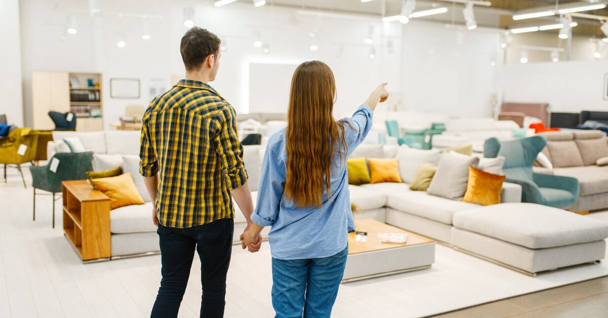 A couple holding hands while shopping for a new couch, standing in a store showroom while the woman points at something in the distance.
