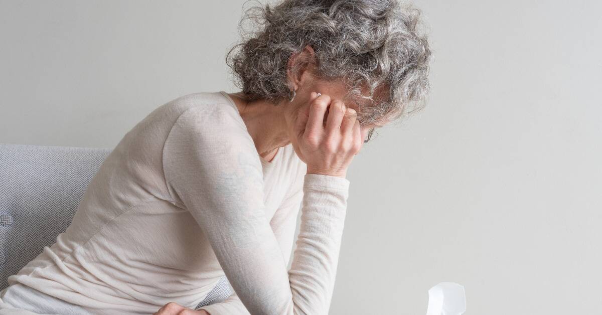 A woman sitting on her bed, obscuring her face with her hand on her cheek, leaning forward with her elbow on her knee.