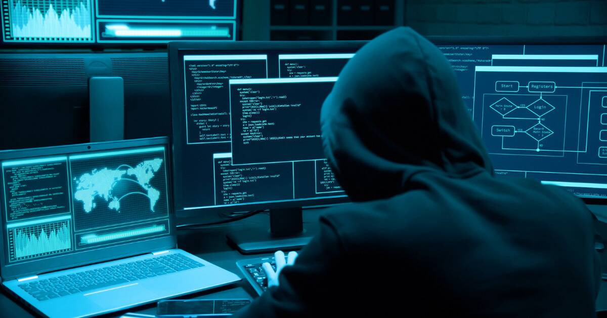 A hacker sat at a computer with many monitors, wearing a black hoodie with the hood up.