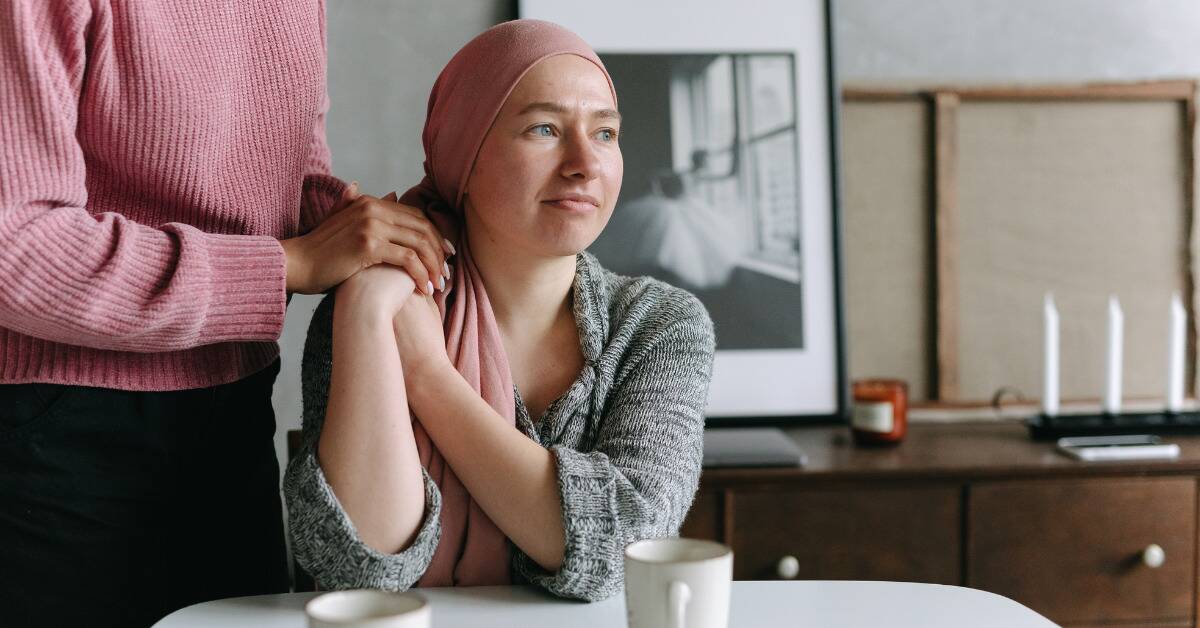 A chemotherapy patient with her head wrapped wits at a table, someone standing next to her, both holding each other's hands at her shoulder.