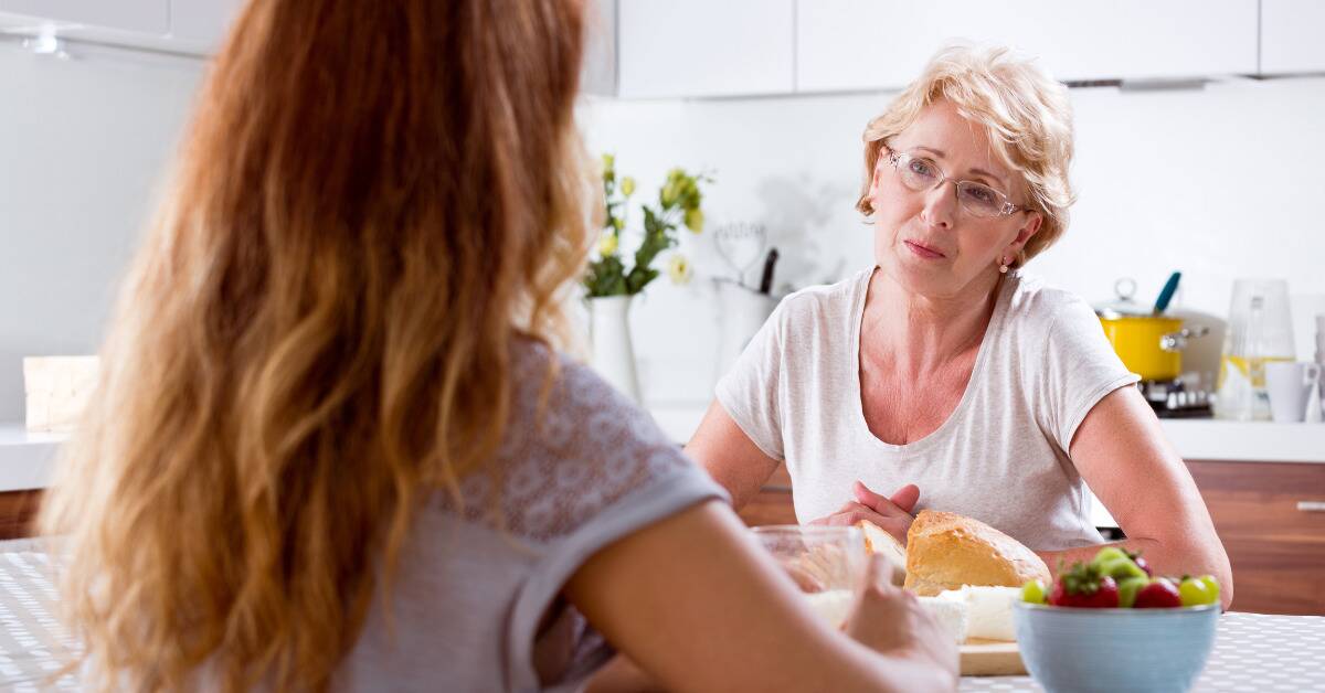 A mother sitting at a dinner table with her daughter, looking disappointed or critical.