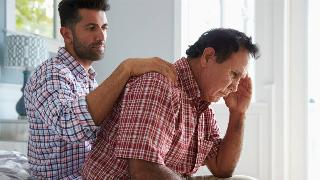 A son sitting behind his father, a comforting hand on his shoulder, as the father leans forward with his hand to his temple in fustration.