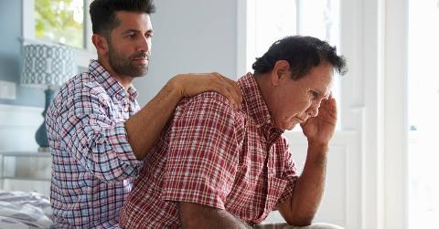 A son sitting behind his father, a comforting hand on his shoulder, as the father leans forward with his hand to his temple in fustration.