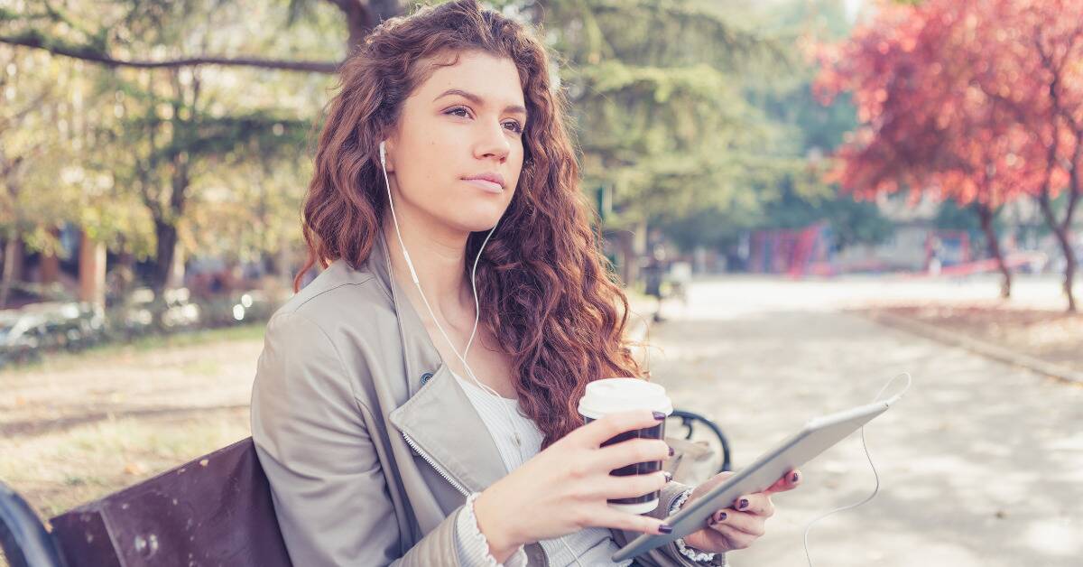 A woman sitting on a park bench, earbuds in and connected to an iPad, a coffee cup in her hand.