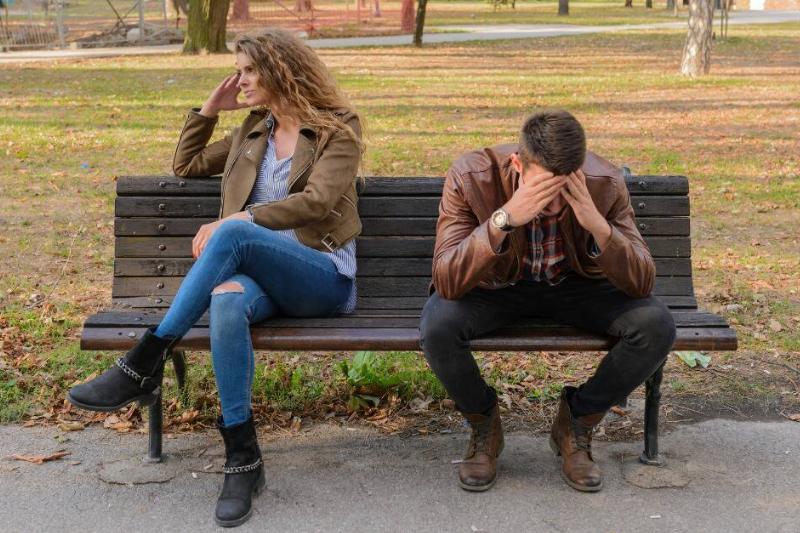 A couple sat on a bench, the woman facing away from the man and looking annoyed while the man has his head in his hands.
