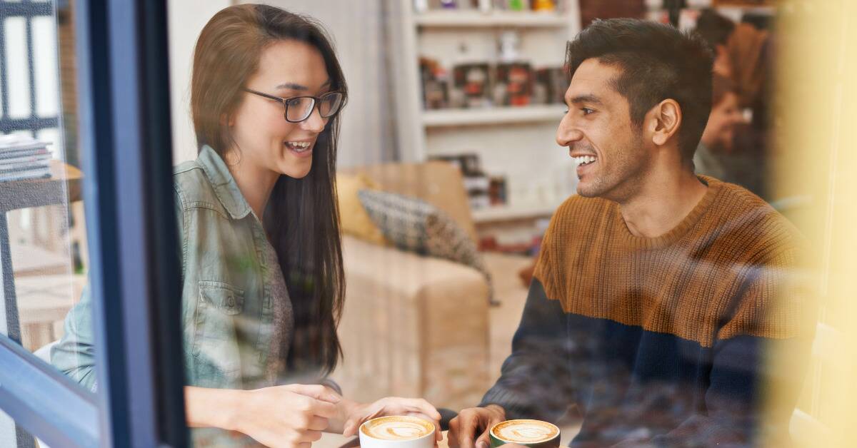 A coupled talking on a cafe date, as seen through a window, both smiling.