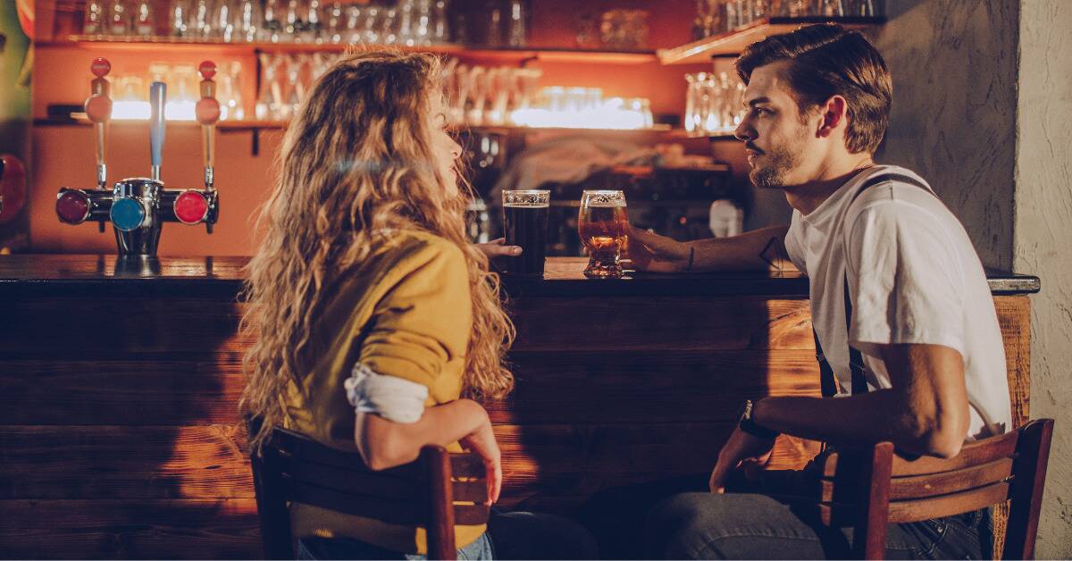 A couple seated next to each other at a bar, angled towards each other, each with a drink as they talk.