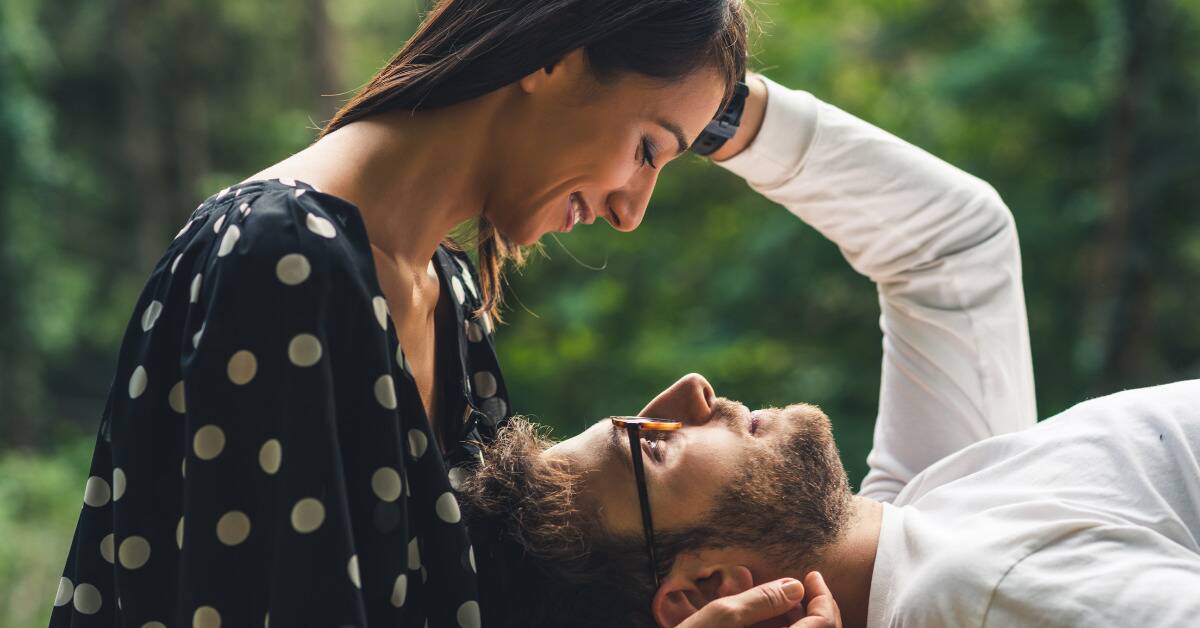 A man laying back, his head in a woman's lap, arm reaching up to the side of her head. She's cradling his head and they're smiling at one another.