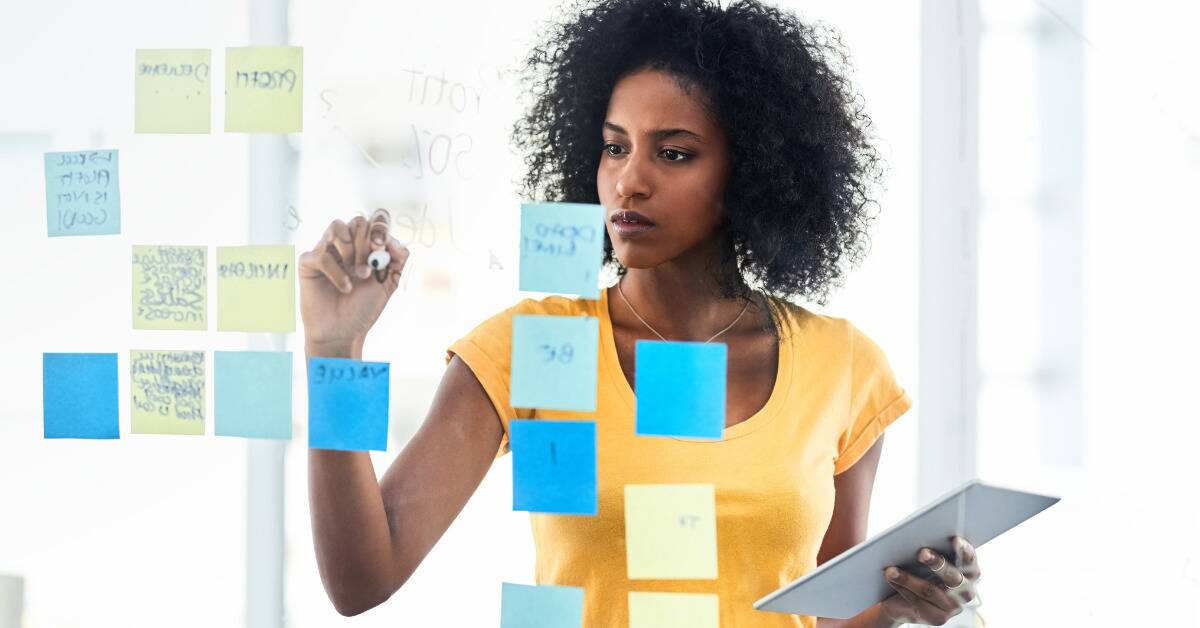 A woman standing in front of a board covered in post-it notes, writing on it with a marker.