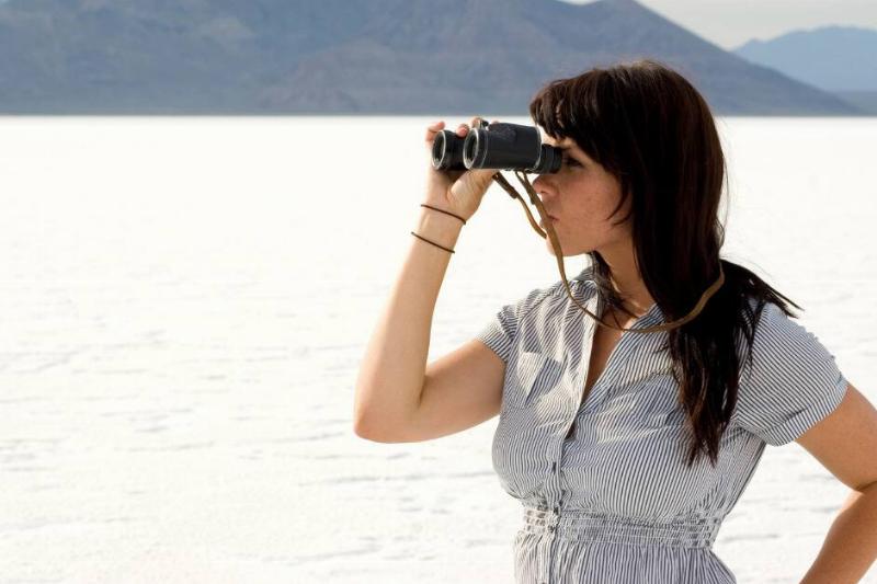 A woman with one hand on her hip, the other holding a pair of binoculars to her eyes, stadning in front of a lake.
