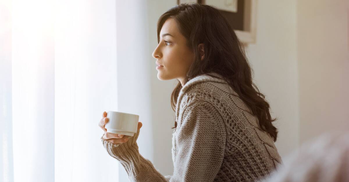 A woman looking wistfully out a window as she holds a cup of tea.