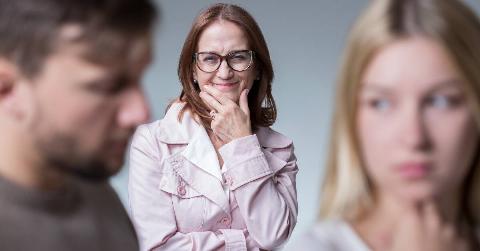 A couple is blurred out in the foreground, the MIL in focus between but behind them, looking scheming.