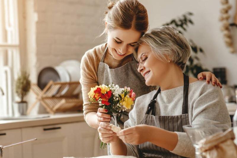 A woman coming up beside her MIL who's sitting baking, handing her a bouquet of flowers.