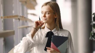 A business woman holding a notebook and pen in her hands smiling as she looks out a window thoughfully.