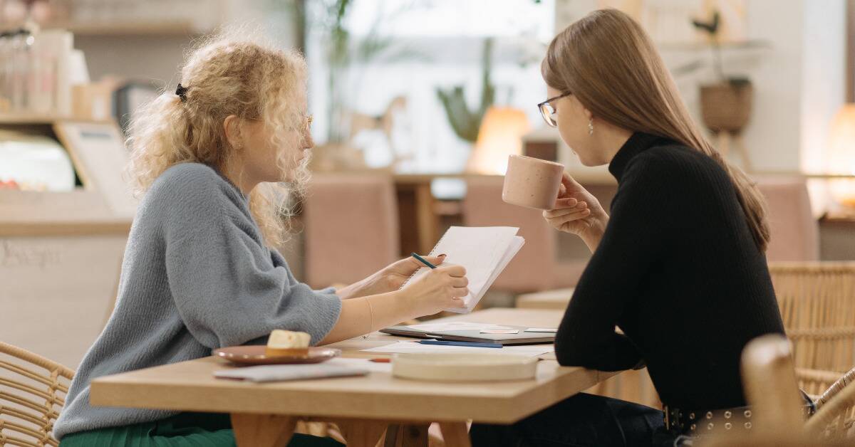Two women at a cafe table, one lifting her mug to have coffee, the other holding up a notepad and showing the other something.