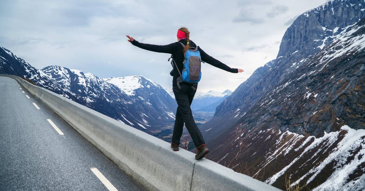 A woman in hiking walking on a barrier on the side of a road next to a mountain range.