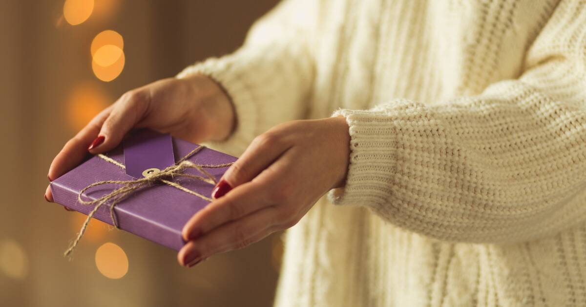 Someone holding a small gift wrapped in purple wrapping paper, holding it out as if giving it to someone.
