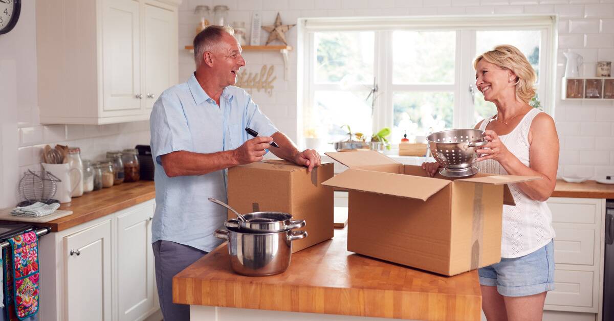 A couple standing in their kitchen, both smiling as they pack up pots and pans.
