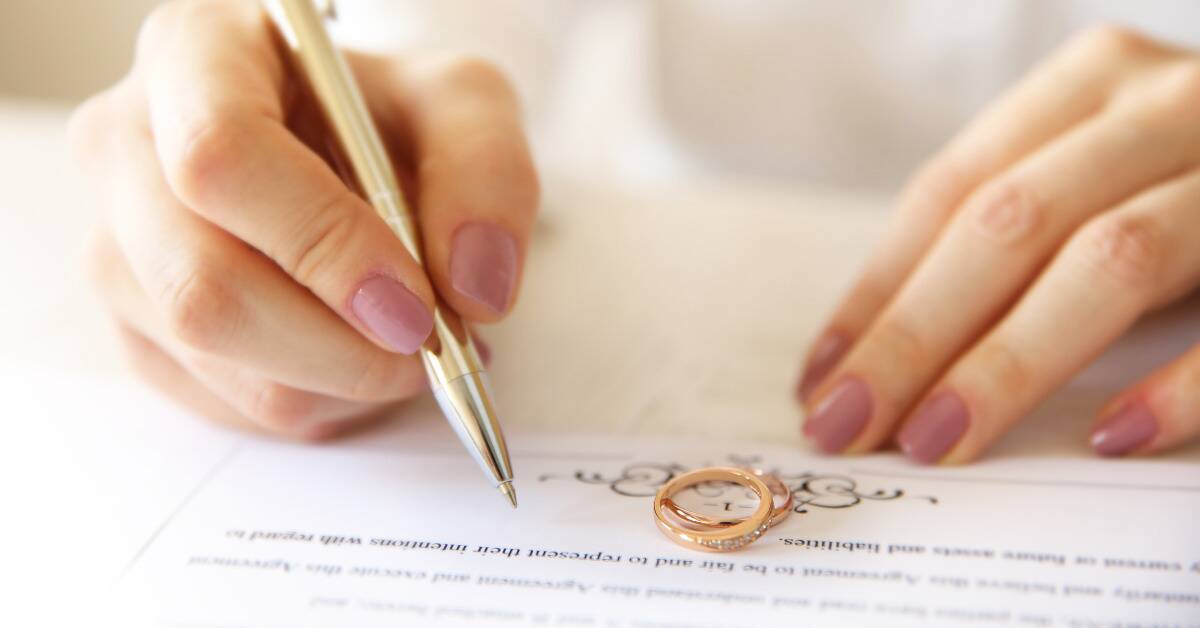 A close shot of a woman with pink painted nails holding a gold pen, signing a marriage document atop which rests her and her spouse's wedding rings.