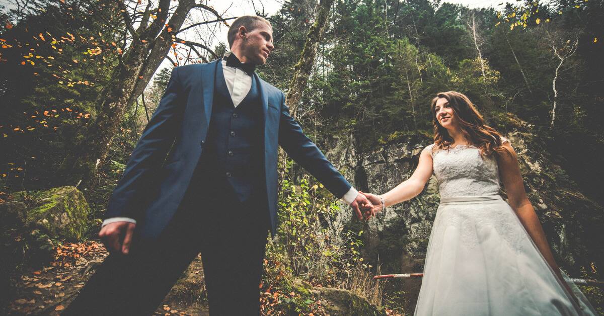 A low-angle shot of a bride and groom holding hands, the groom leading the bride through a forest path.