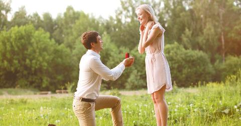 A young couple in a field, the man on one knee proposing to the woman, both smiling and looking excited.
