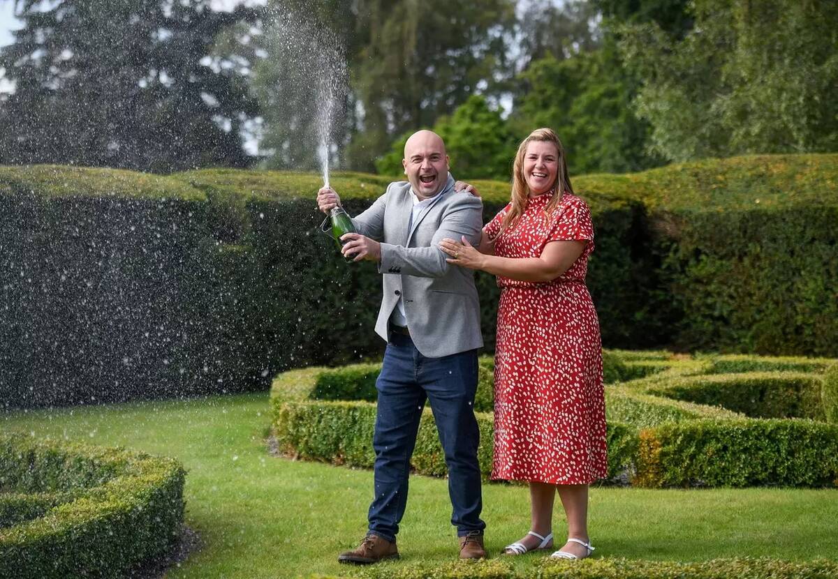 Kathleen and Mark Reece outside for a photoshoot, Mark popping a bottle of champagne as they both laugh.