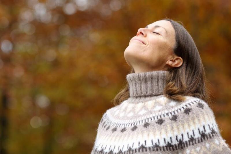 A woman in a knit sweater outside during autumn, her eyes closed, head tilted up to the sky as she smiles.