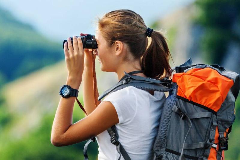 A woman with a hiking backpack looking through a pair of binoculars while outside.