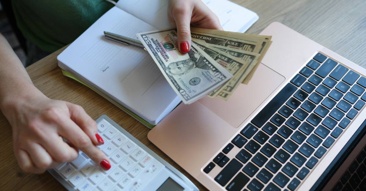 A woman holding a fanned out stack of cash while punching numbers into her calculator.