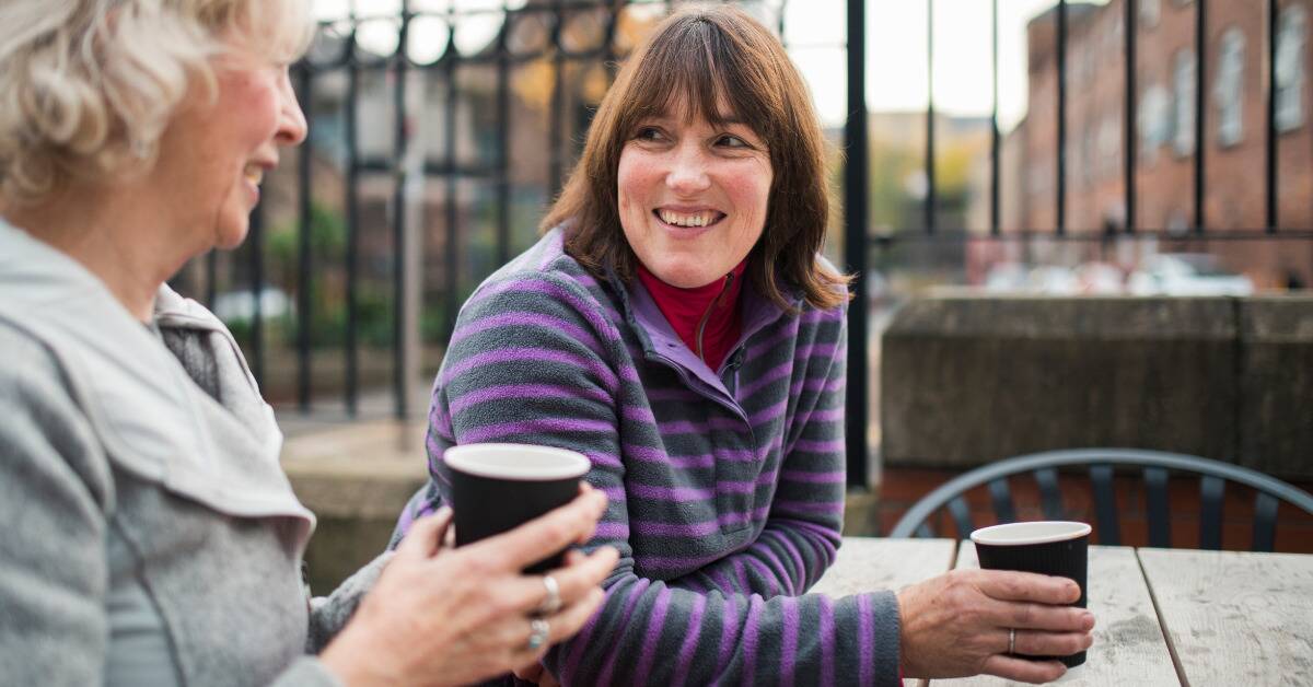 Two women talk over cups of coffee, sitting outside, both smiling.