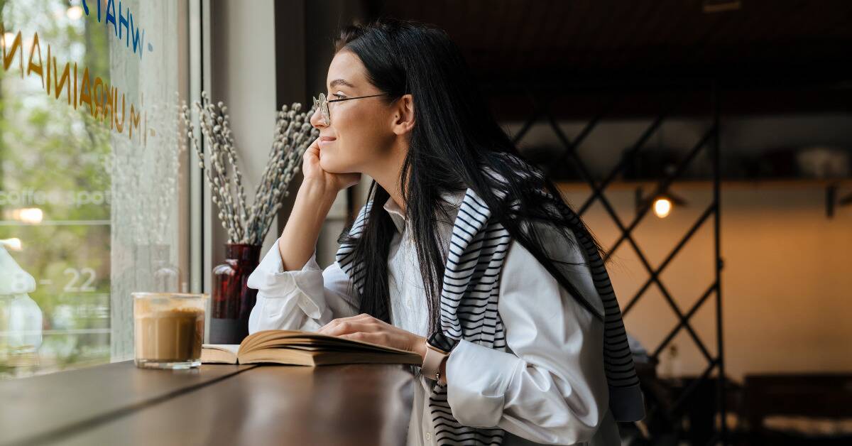 A woman sitting at a cafe with a  book, looking out the window wistfully.